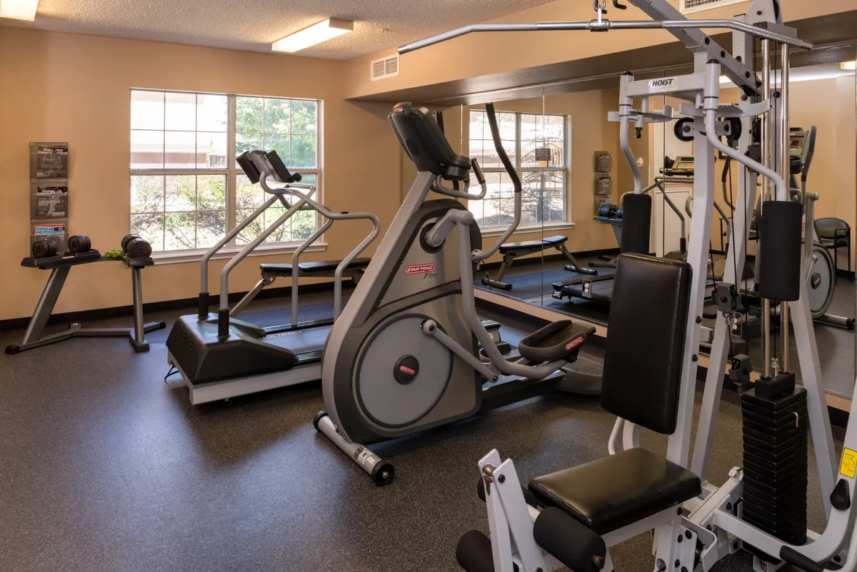 A bright, contemporary fitness center with large windows, featuring an elliptical machine, stationary bike, weight machine, and space for group fitness. The area is designed for energizing workouts and self-improvement.