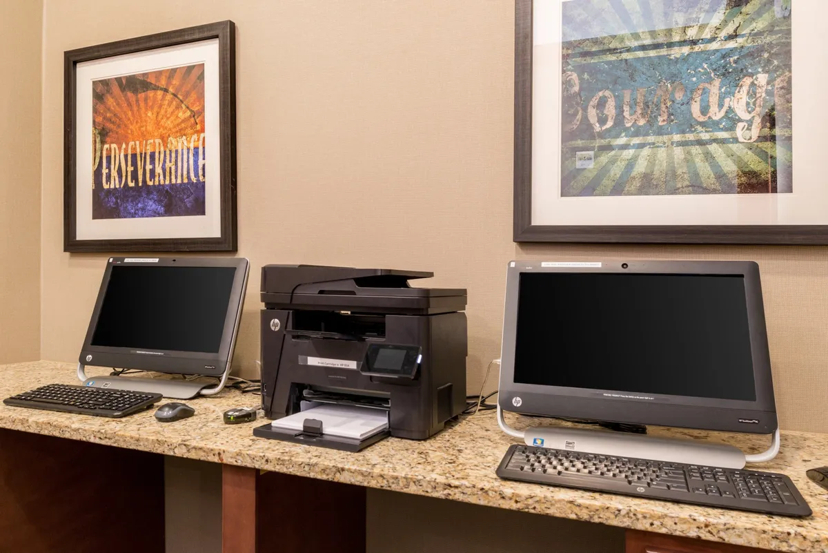 This image depicts a well-lit workspace featuring two desktop computers, a printer, and framed inspirational artwork. The modern setup suggests a productive environment, well-suited for both business and leisure travelers who need to work remotely or catch up on tasks.
