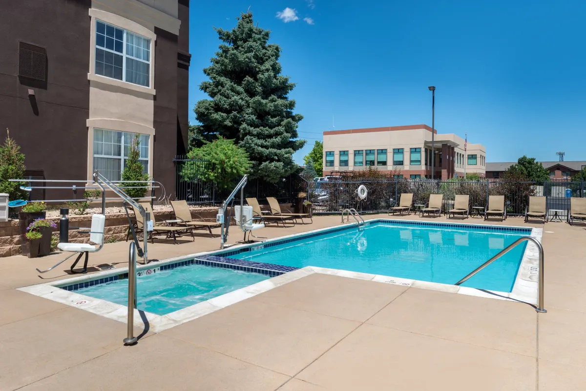 An inviting exterior view of the building facade with a shimmering swimming pool surrounded by lounge chairs and lush landscaping. The scene captures a leisurely atmosphere with the pool as the focal point, offering ample space for relaxation and casual socializing.