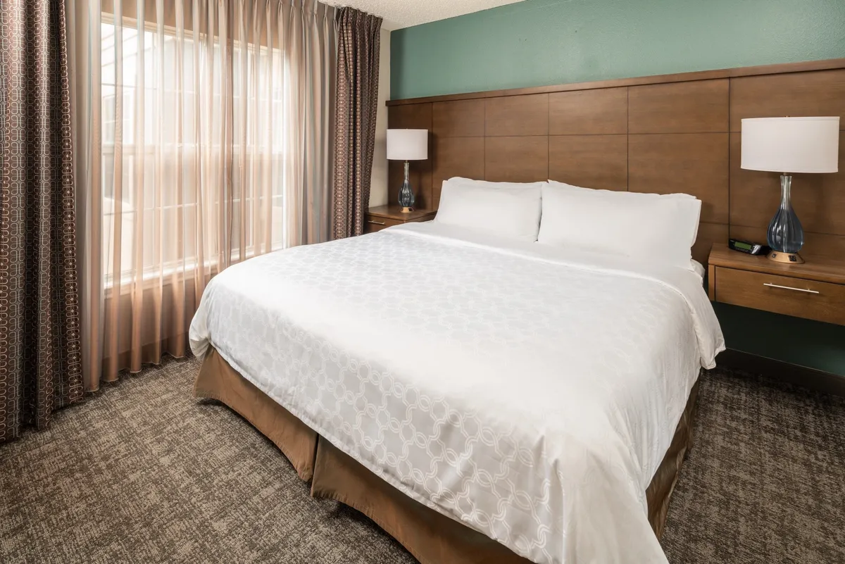 A tranquil bedroom scene with a king-size bed adorned in luxury linens, soft sunlight streaming through curtains, and elegant nightstands with lamps. The setup is ideal for reading, unwinding, or waking up refreshed. The overall ambiance is serene and inviting, perfectly suited to restful sleep and relaxation.